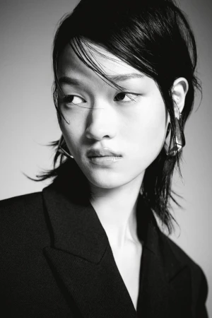 Monochrome portrait of an Asian female model with dark hair wearing a black blazer and geometric earrings against a grey backdrop.
