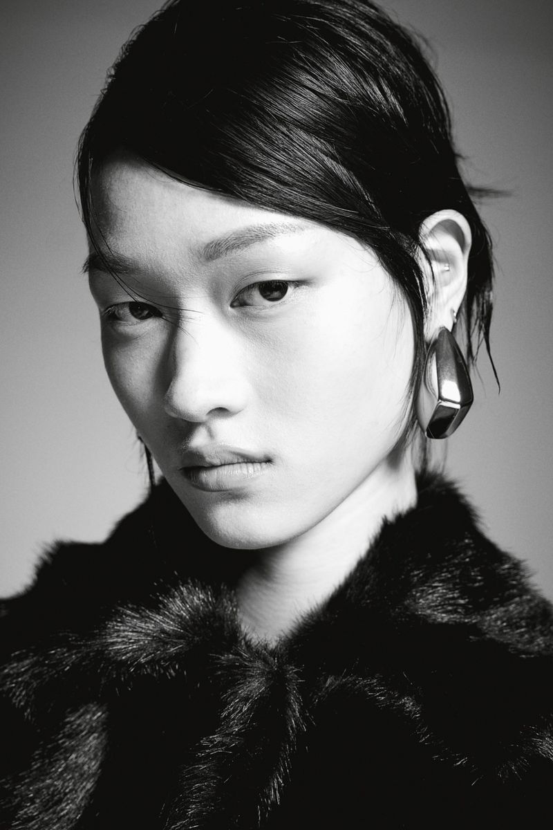 Close-up black and white portrait of a model wearing a textured faux fur coat and sculptural metallic earrings against a grey backdrop
