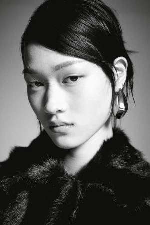 Close-up black and white portrait of a model wearing a textured faux fur coat and sculptural metallic earrings against a grey backdrop