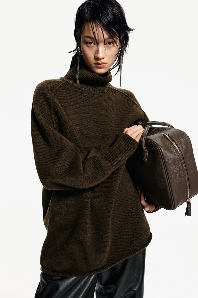 A woman poses in a studio wearing a green turtleneck sweater, black pants, and holding a brown leather handbag with white studio background.