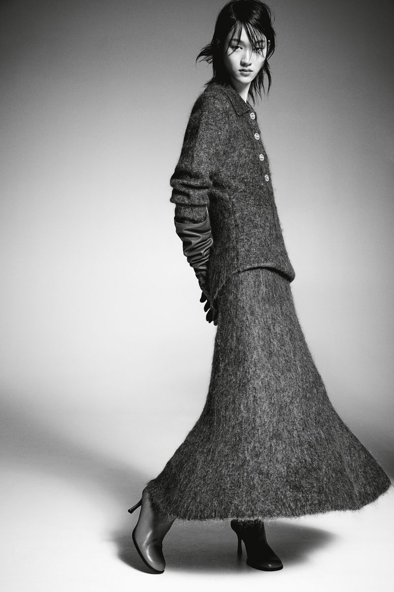 A woman models a gray textured two-piece suit with long sleeves and a long skirt, accessorized with gloves and high heels in a studio shot.