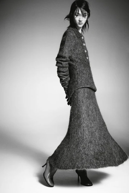 A woman models a gray textured two-piece suit with long sleeves and a long skirt, accessorized with gloves and high heels in a studio shot.