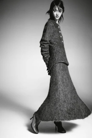 A woman models a gray textured two-piece suit with long sleeves and a long skirt, accessorized with gloves and high heels in a studio shot.