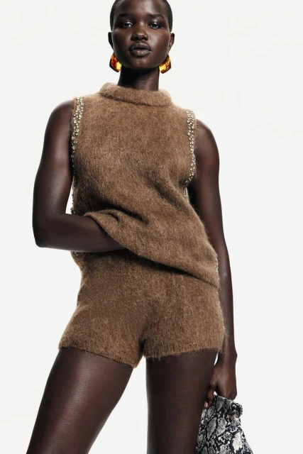 Fashion shot of a Black female model wearing a brown sweater and shorts set with amber earrings, against a bright white background.