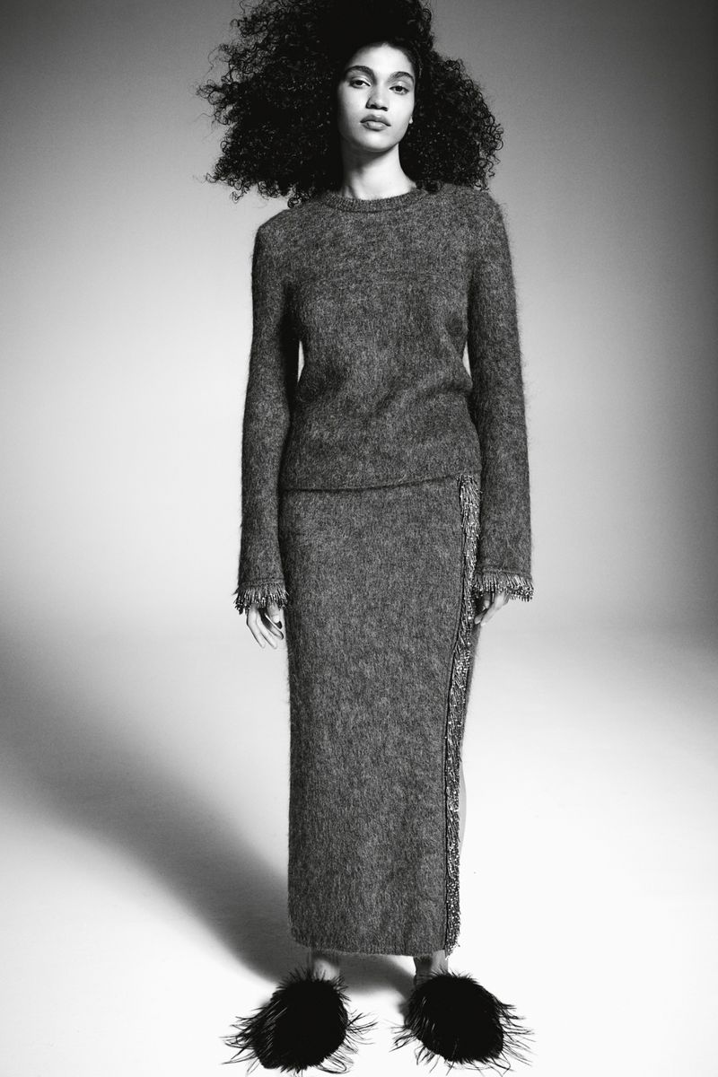 Model with voluminous curly hair wearing a textured grey knit sweater and skirt against a seamless white background.