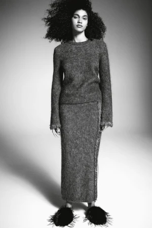 Model with voluminous curly hair wearing a textured grey knit sweater and skirt against a seamless white background.