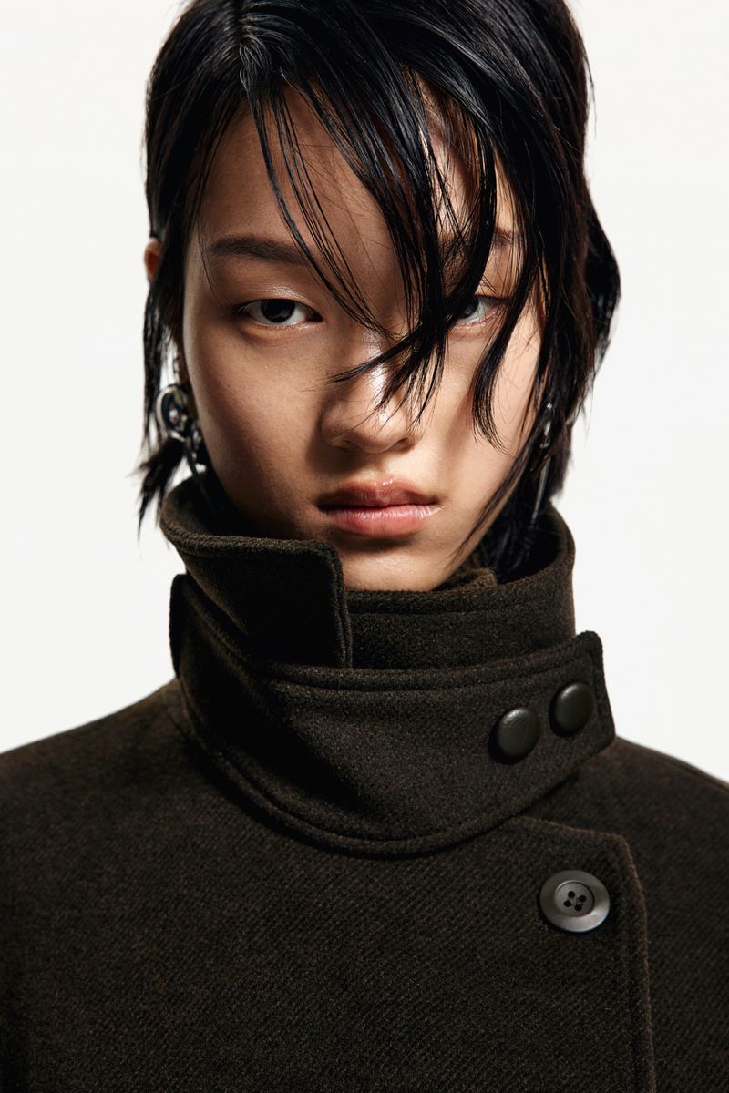 A young Asian woman wearing a brown coat with a high collar.  The photo is a close up shot with a white background.