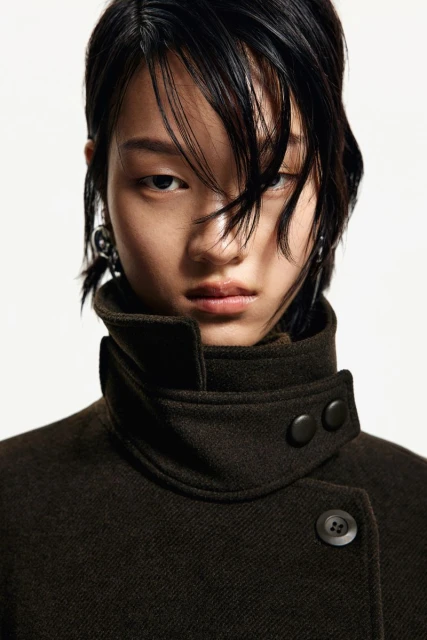 A young Asian woman wearing a brown coat with a high collar.  The photo is a close up shot with a white background.