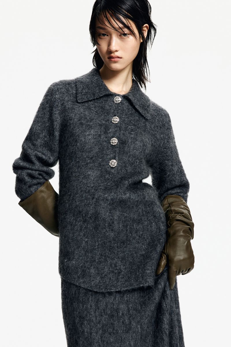 An Asian woman poses in a studio wearing a grey sweater dress with crystal buttons and long green leather gloves against a white backdrop.
