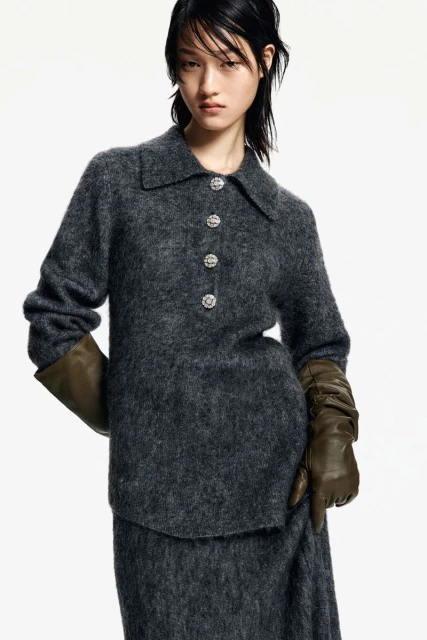 An Asian woman poses in a studio wearing a grey sweater dress with crystal buttons and long green leather gloves against a white backdrop.