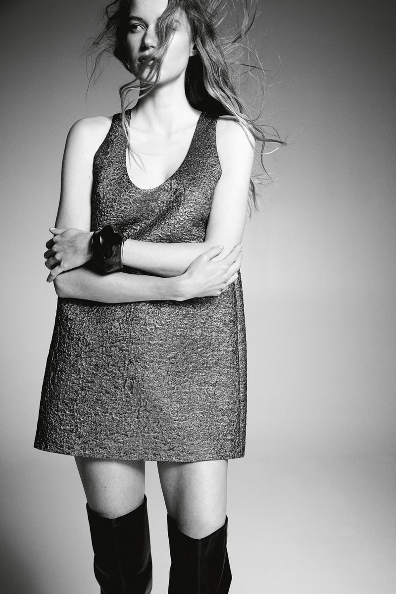 Monochrome portrait of a young woman in a mini dress and boots with wind-swept hair, posing in a studio environment with simple backdrop.