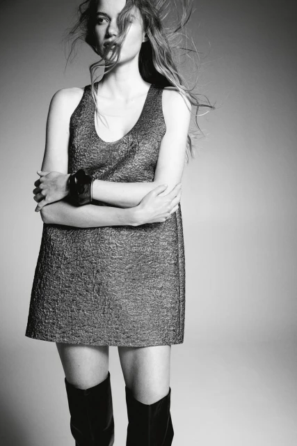Monochrome portrait of a young woman in a mini dress and boots with wind-swept hair, posing in a studio environment with simple backdrop.