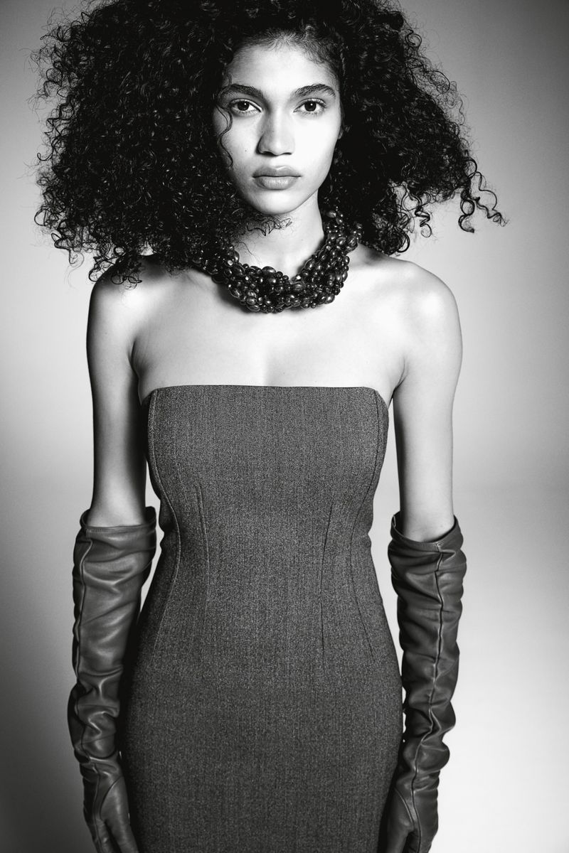 Model wearing a strapless dark dress and leather gloves with a beaded necklace against a plain grey studio backdrop