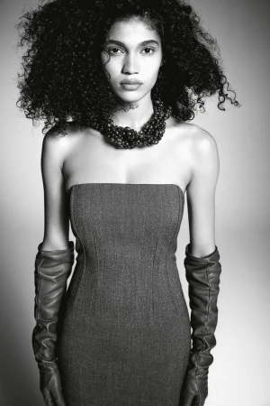 Model wearing a strapless dark dress and leather gloves with a beaded necklace against a plain grey studio backdrop
