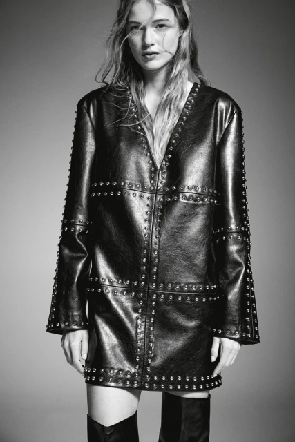 Studio shot of a woman in a studded black leather jacket dress and knee-high boots, showcasing a high-fashion editorial aesthetic.