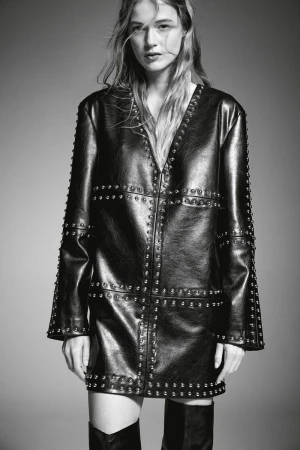 Studio shot of a woman in a studded black leather jacket dress and knee-high boots, showcasing a high-fashion editorial aesthetic.