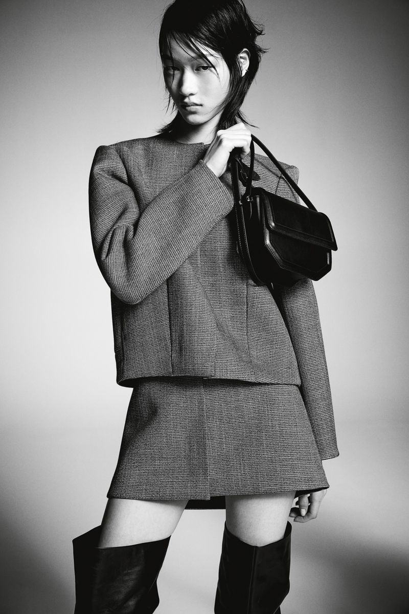 Monochrome studio shot of a stylish woman with short hair in a grey skirt suit, accessorized with black leather boots and a handbag.