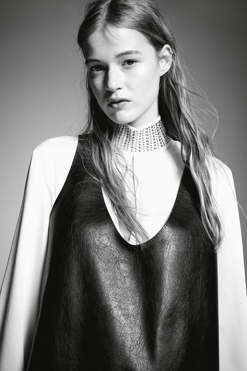 Portrait of a young woman in a black dress over a white long-sleeved shirt, posing against a grey background, with a medium shot.