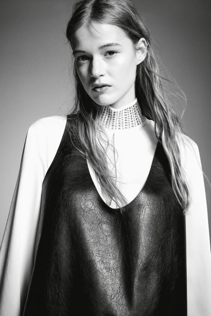 Portrait of a young woman in a black dress over a white long-sleeved shirt, posing against a grey background, with a medium shot.