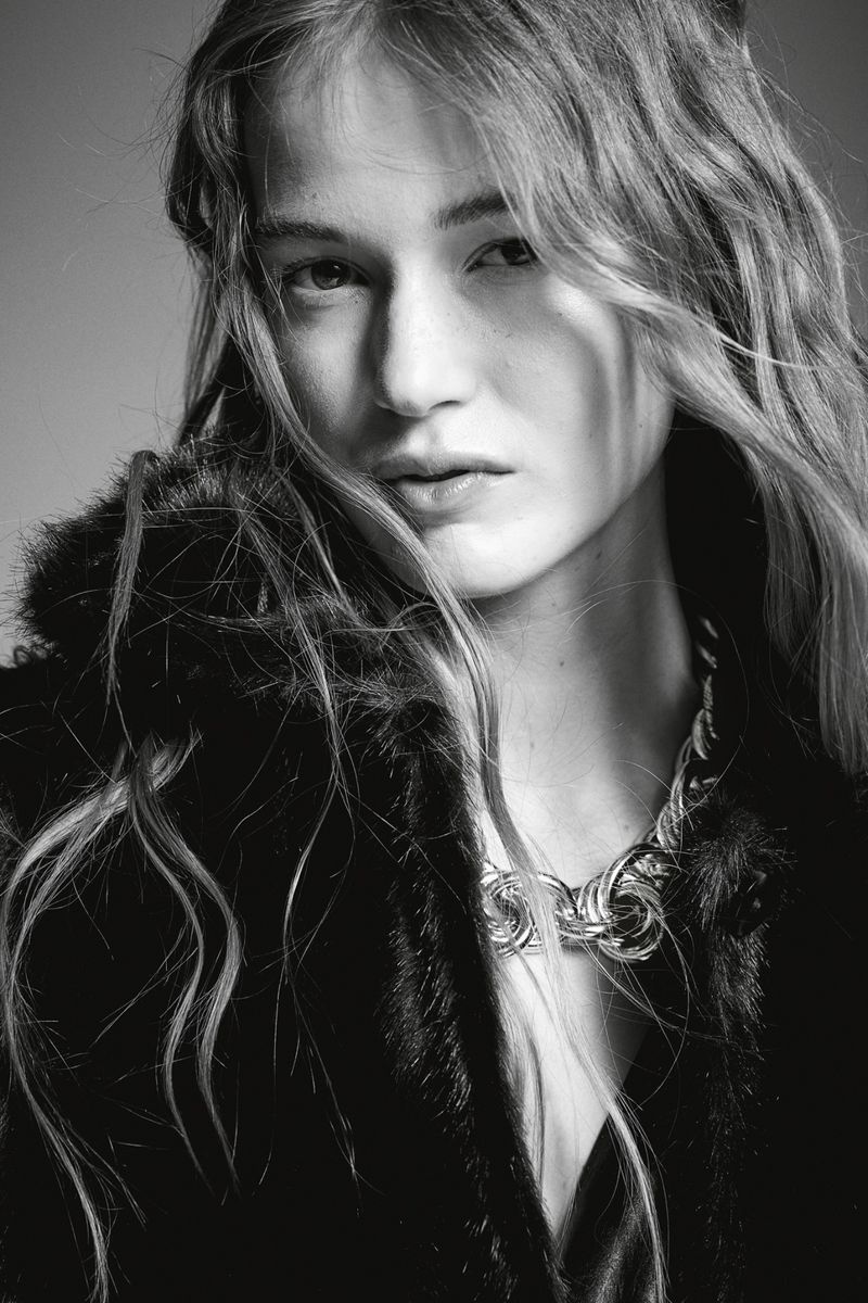 Close-up portrait of a woman with long wavy hair wearing a textured dark jacket and a chunky silver chain necklace against a grey backdrop.