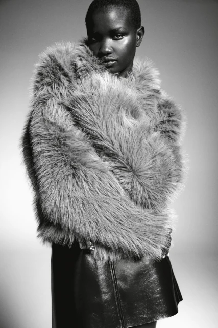 Monochrome portrait of a Black woman with short hair wearing a furry grey coat and black skirt against a neutral background.