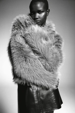 Monochrome portrait of a Black woman with short hair wearing a furry grey coat and black skirt against a neutral background.