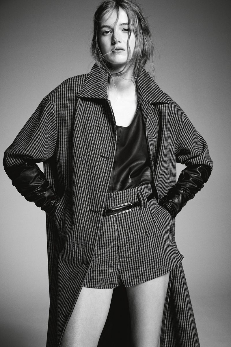 Black-and-white portrait of a female model wearing a plaid coat, leather top, and matching shorts, posing in a studio setting.