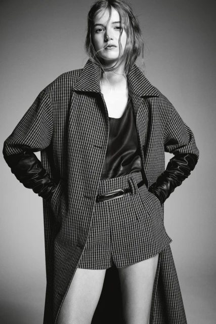 Black-and-white portrait of a female model wearing a plaid coat, leather top, and matching shorts, posing in a studio setting.