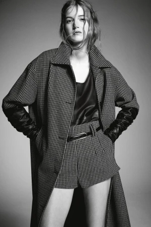 Black-and-white portrait of a female model wearing a plaid coat, leather top, and matching shorts, posing in a studio setting.