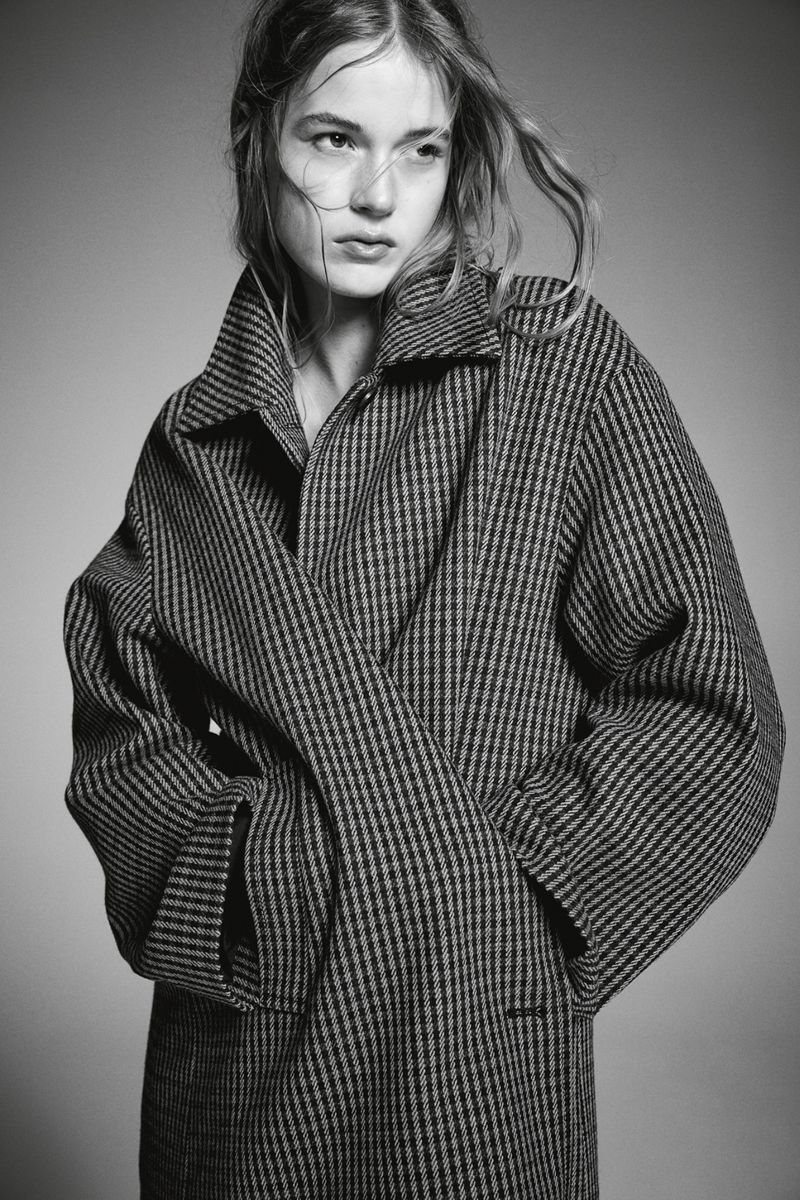 Monochrome portrait of a young fair-skinned woman with light hair wearing a striped wool coat, posing in a neutral indoor setting.