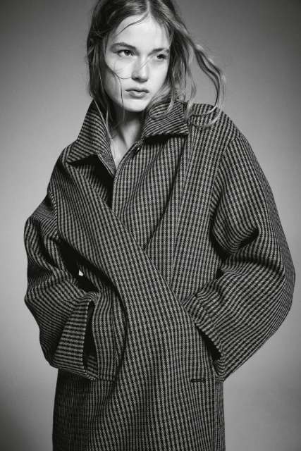 Monochrome portrait of a young fair-skinned woman with light hair wearing a striped wool coat, posing in a neutral indoor setting.