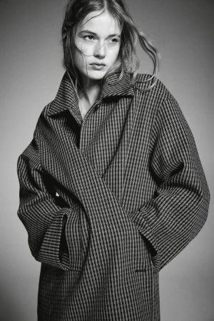 Monochrome portrait of a young fair-skinned woman with light hair wearing a striped wool coat, posing in a neutral indoor setting.