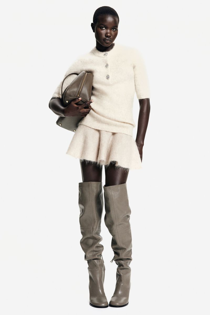 Full-length shot of a Black female fashion model in a short-sleeved sweater, mini skirt, knee-high boots, and a shoulder bag against a white background.