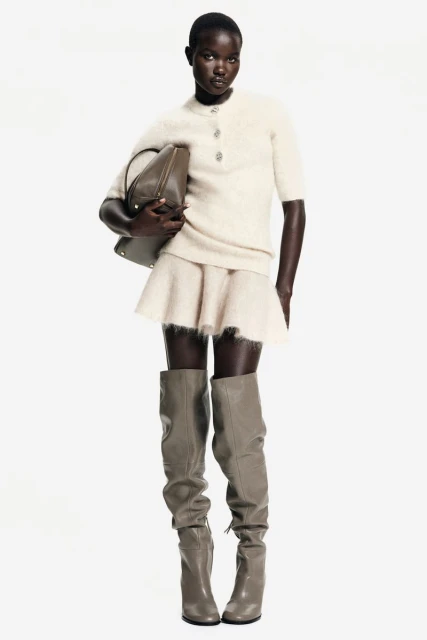 Full-length shot of a Black female fashion model in a short-sleeved sweater, mini skirt, knee-high boots, and a shoulder bag against a white background.