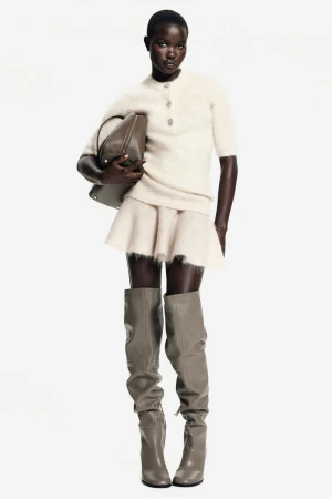 Full-length shot of a Black female fashion model in a short-sleeved sweater, mini skirt, knee-high boots, and a shoulder bag against a white background.