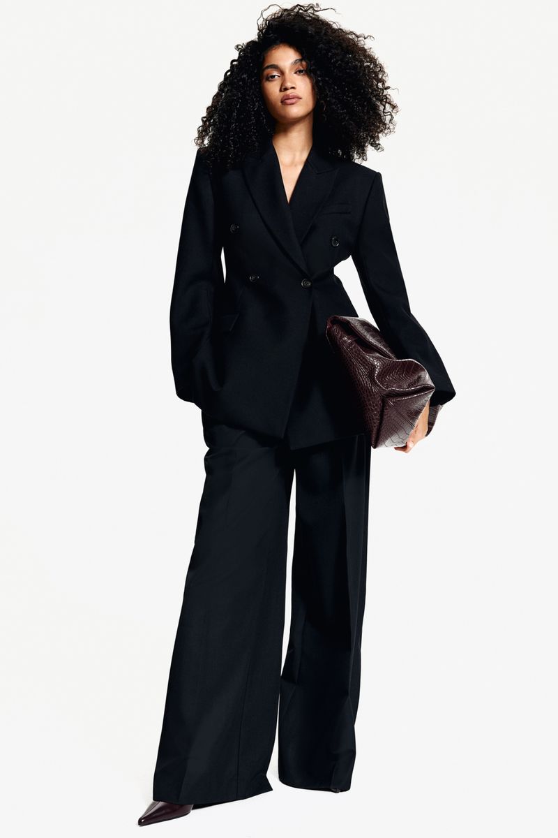Fashionable adult female stands in a photo studio wearing a dark pantsuit with a handbag. She poses with a neutral facial expression.