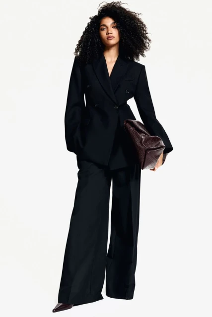 Fashionable adult female stands in a photo studio wearing a dark pantsuit with a handbag. She poses with a neutral facial expression.