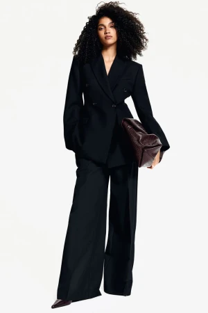 Fashionable adult female stands in a photo studio wearing a dark pantsuit with a handbag. She poses with a neutral facial expression.