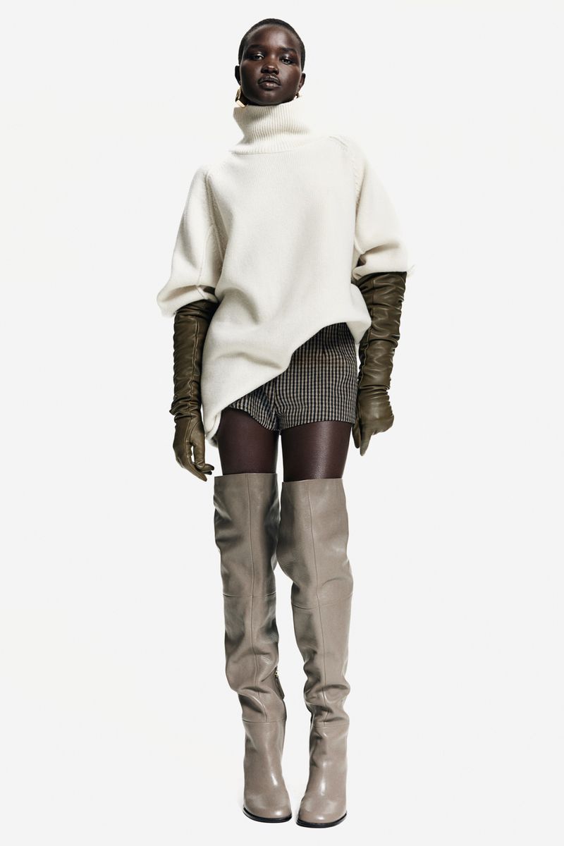Fashion shot of a Black female model in a turtleneck sweater, gloves, shorts, and thigh-high boots, posing in a white studio.