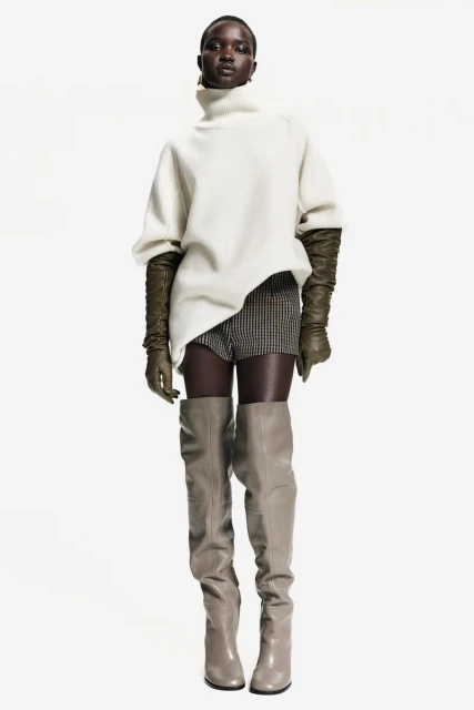 Fashion shot of a Black female model in a turtleneck sweater, gloves, shorts, and thigh-high boots, posing in a white studio.