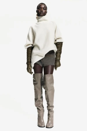 Fashion shot of a Black female model in a turtleneck sweater, gloves, shorts, and thigh-high boots, posing in a white studio.