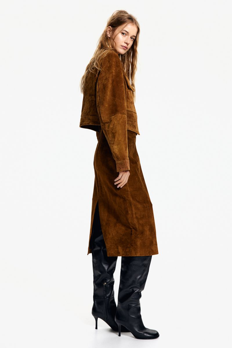 A fair-skinned woman in a brown suede jacket and skirt with black high-heeled boots, shot in a full shot against a white backdrop.