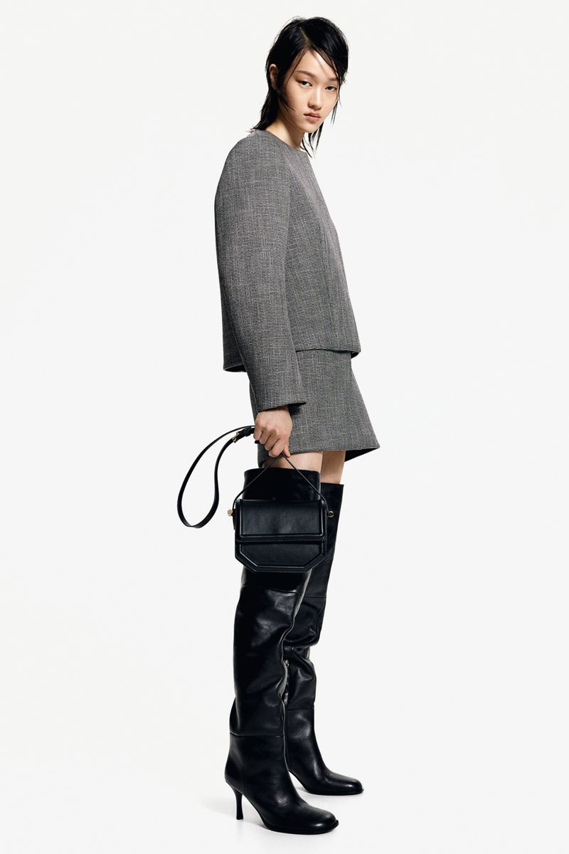 A woman in a gray tweed suit and black thigh-high heeled boots poses with a black purse against a white backdrop.