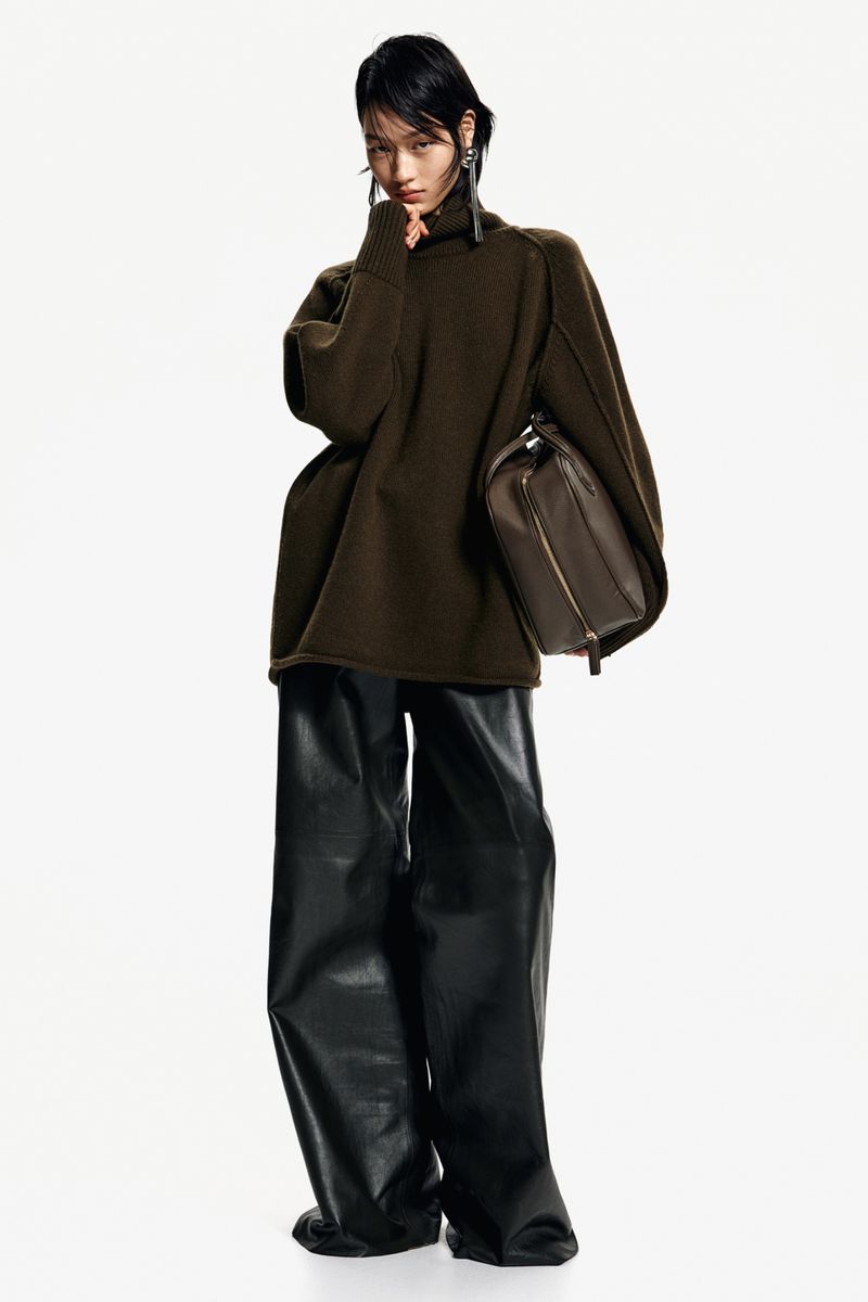 A stylish Asian female model poses in a studio wearing an olive green sweater, black leather pants, and carrying a brown leather handbag.