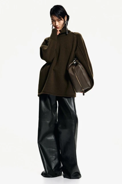 A stylish Asian female model poses in a studio wearing an olive green sweater, black leather pants, and carrying a brown leather handbag.