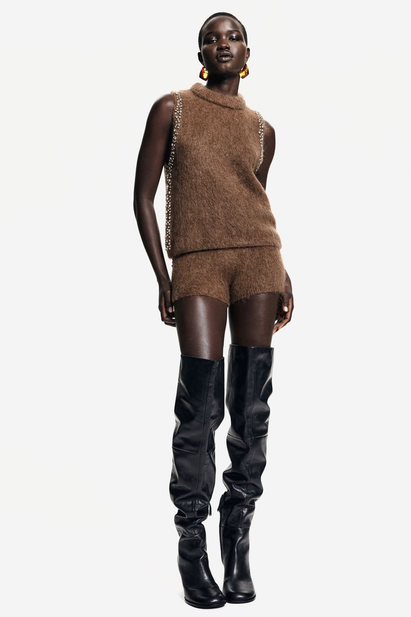 Full-length shot of a Black female fashion model in a brown knit top and shorts with black thigh-high boots, posing against a white backdrop.