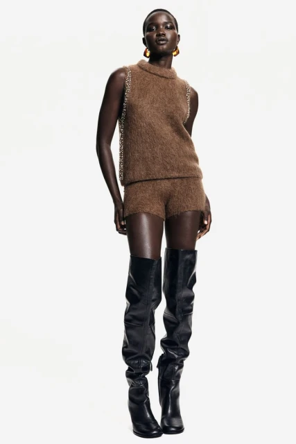 Full-length shot of a Black female fashion model in a brown knit top and shorts with black thigh-high boots, posing against a white backdrop.