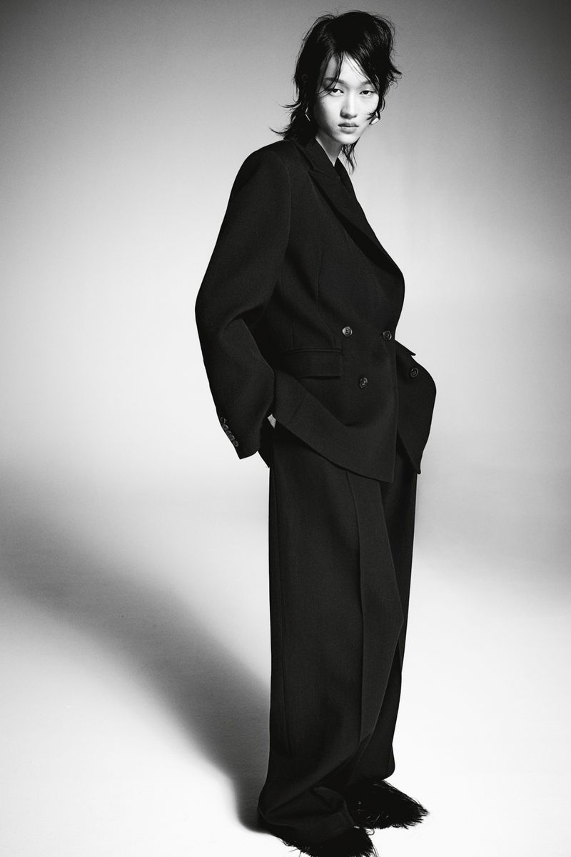 Monochrome portrait of a stylish young woman in an oversized black suit and wide-leg trousers against a neutral studio backdrop.