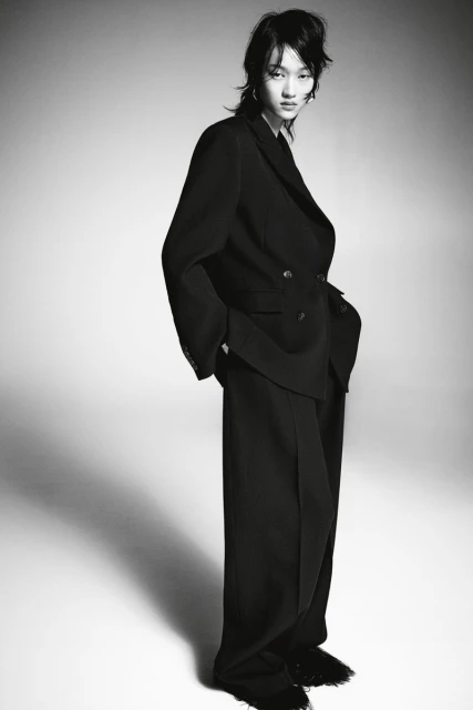 Monochrome portrait of a stylish young woman in an oversized black suit and wide-leg trousers against a neutral studio backdrop.