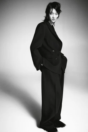 Monochrome portrait of a stylish young woman in an oversized black suit and wide-leg trousers against a neutral studio backdrop.